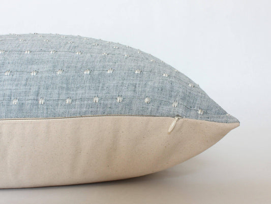 Close-up of blue textured pillow with white stitched dots and beige fabric bottom with zipper, on white background. Come see us at Awaken, Franklin, Tennessee