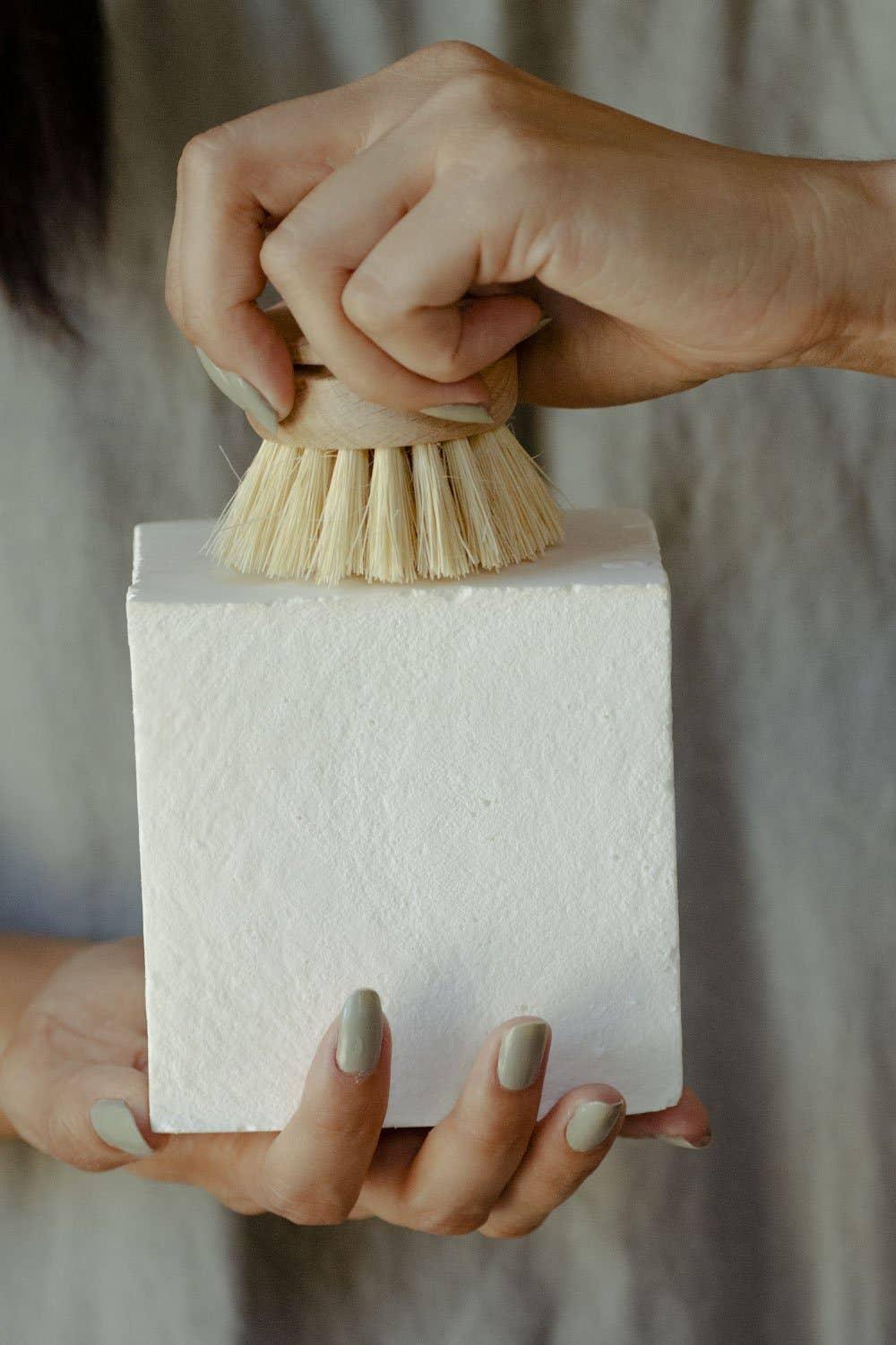 Hands with light green nail polish holding a wooden scrub brush and a white block. Come see us at Awaken, Franklin, Tennessee