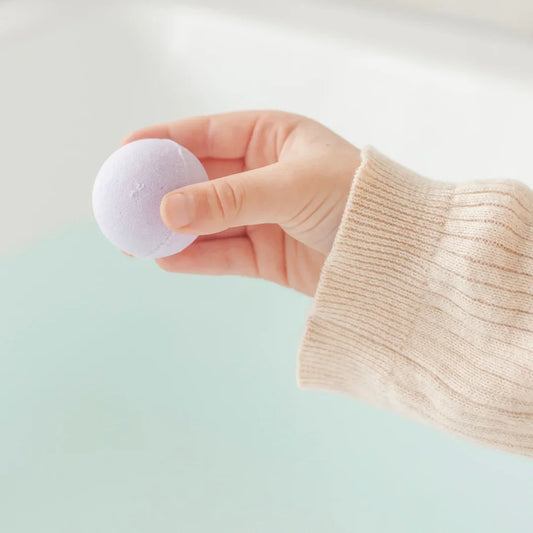 Hand holding a lavender bath bomb over a water-filled tub, wearing a beige sweater. Come see us at Awaken, Franklin, Tennessee