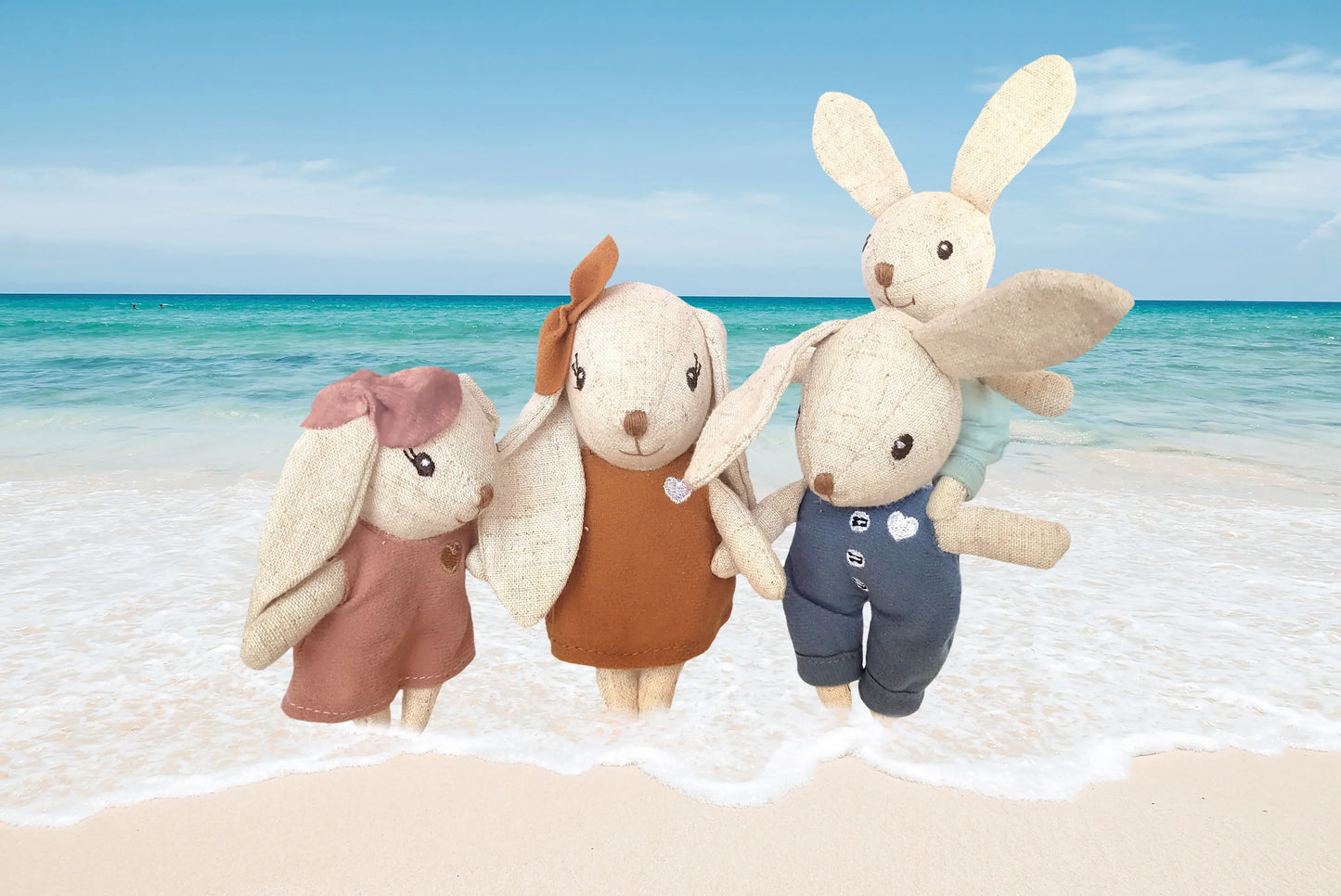 Four handmade cloth bunny dolls on sandy beach with ocean waves, bright sky. Come see us at Awaken, Franklin, Tennessee