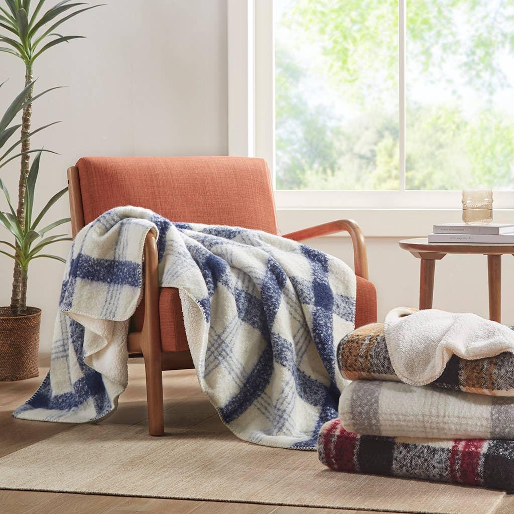 Cozy plaid sherpa blankets on orange chair and stacked nearby in bright room with plant, window. Come see us at Awaken, Franklin, Tennessee