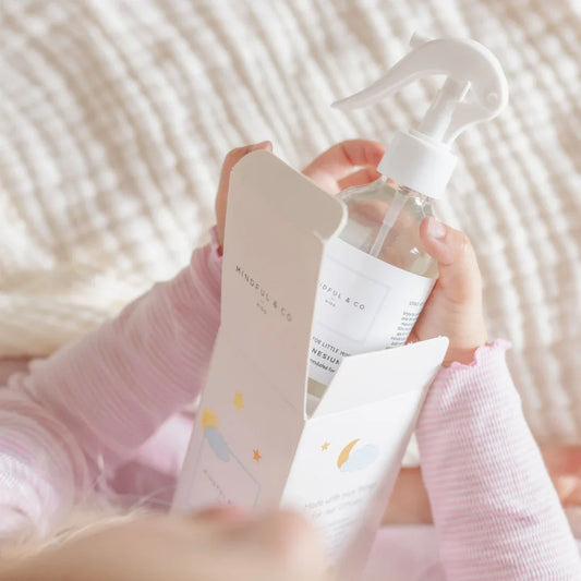 Child in pink pajamas holding Mindful & Co kids spray bottle and box on cozy blanket, Come see us at Awaken, Franklin, Tennessee