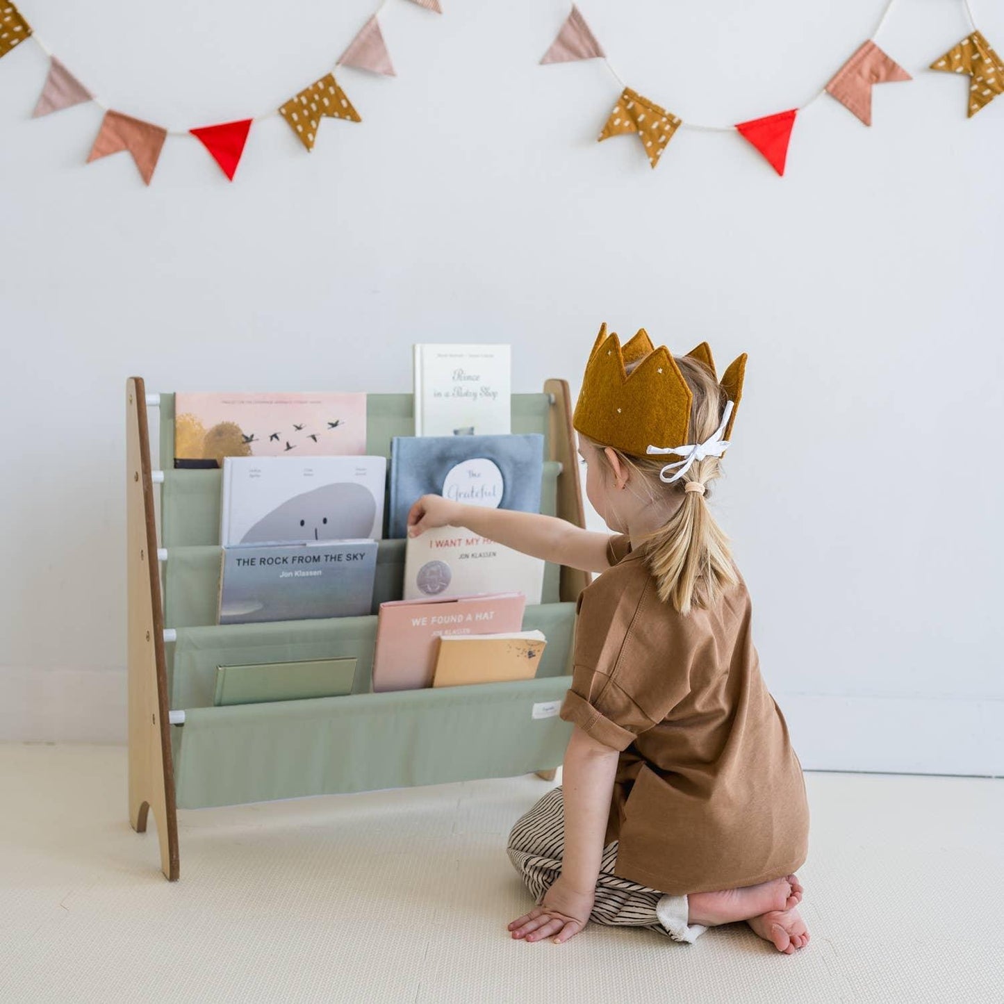 Child wearing a felt crown reading books from a wooden rack in a decorated room, Come see us at Awaken, Franklin, Tennessee