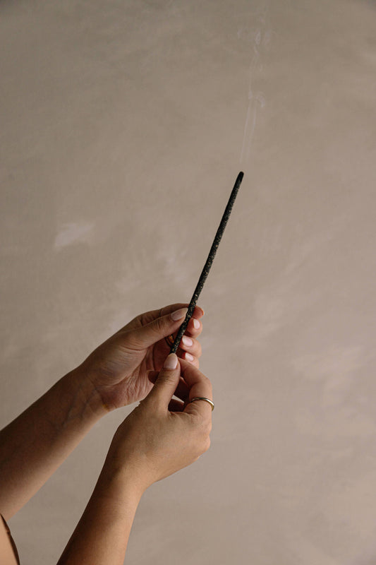 Hands holding a burning black incense stick with smoke against a neutral background. Come see us at Awaken, Franklin, Tennessee