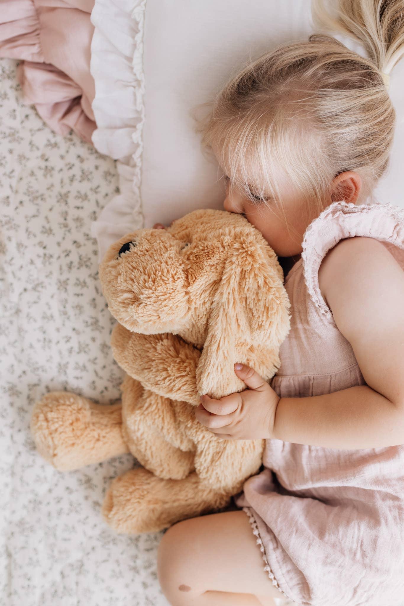 Toddler girl in pink dress cuddling a plush dog toy on floral bedding Come see us at Awaken, Franklin, Tennessee