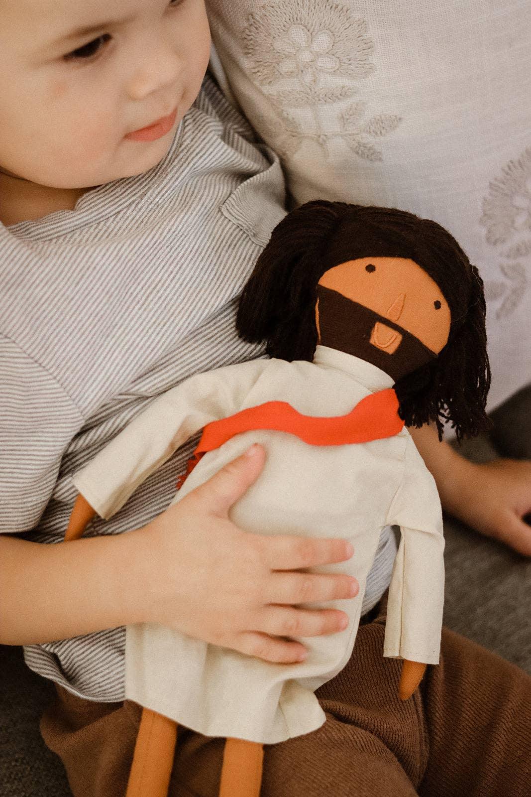 Toddler holding a soft Jesus doll with a red scarf on a couch, cozy setting. Come see us at Awaken, Franklin, Tennessee