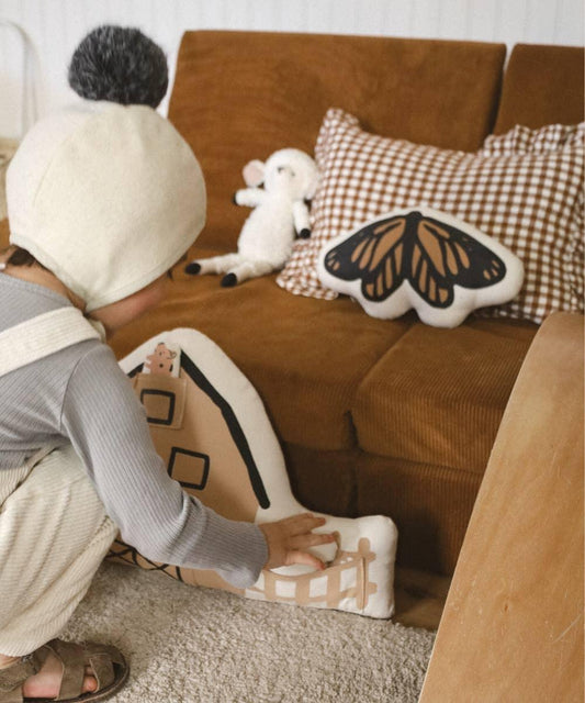 Child playing with soft house-shaped toy near brown couch with butterfly and gingham pillows. Come see us at Awaken, Franklin, Tennessee