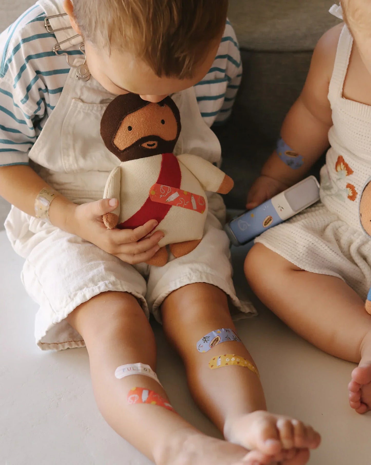 Toddler holding plush Jesus doll, colorful bandages on legs, sitting with baby holding a device. Come see us at Awaken, Franklin, Tennessee