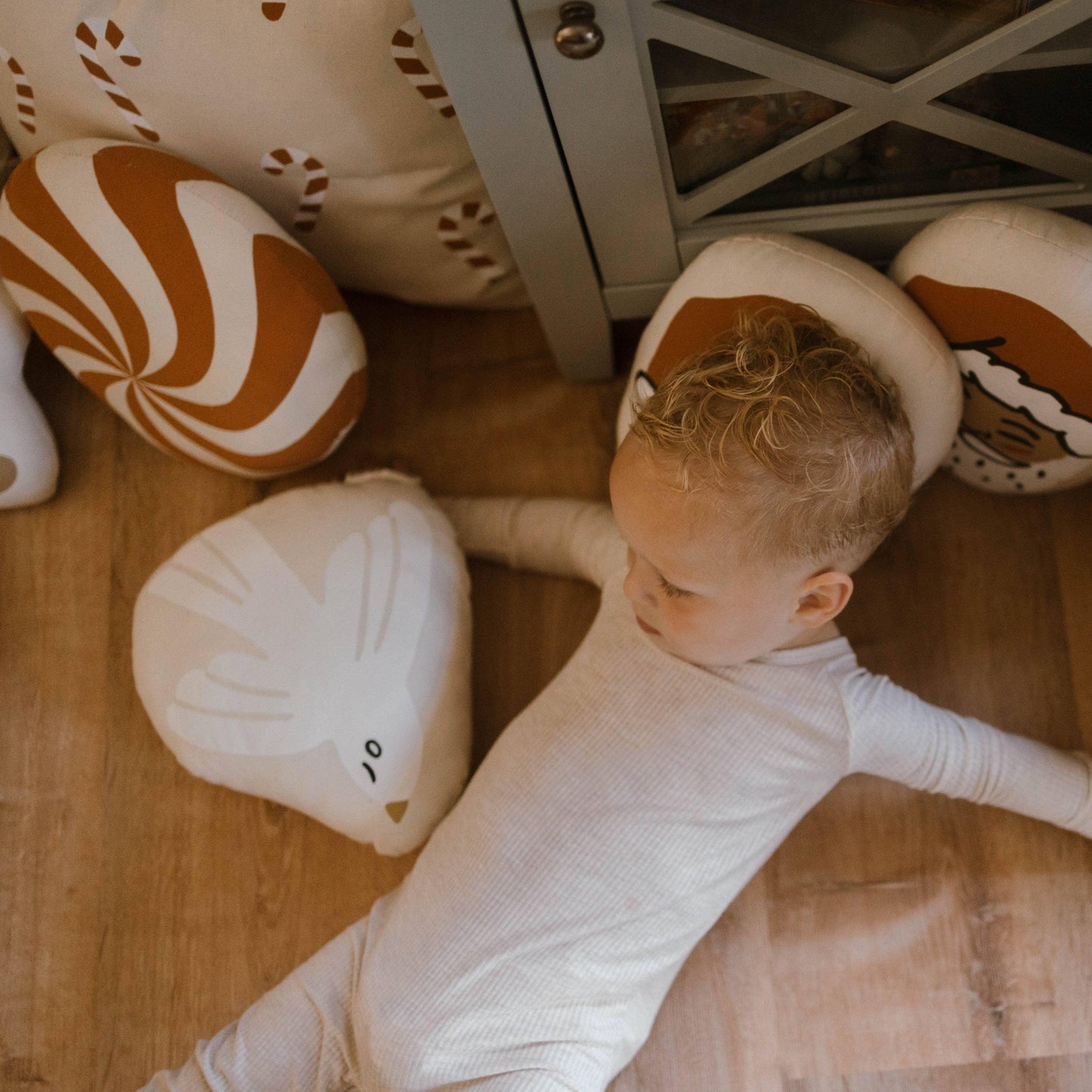Toddler in white pajamas lying on wooden floor with bird and candy-themed pillows. Come see us at Awaken, Franklin, Tennessee