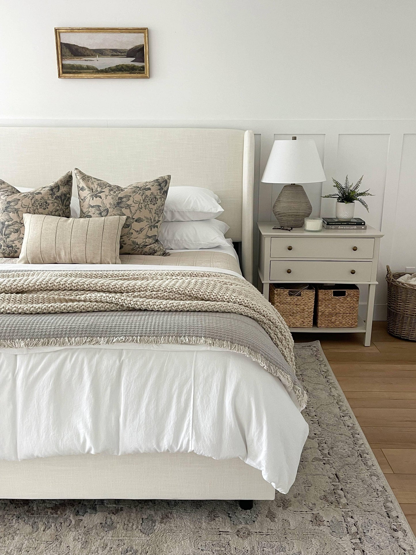 Cozy bedroom with beige upholstered bed, floral pillows, knit blanket, nightstand with lamp and decor. Come see us at Awaken, Franklin, Tennessee