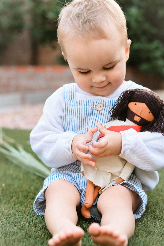 Toddler in striped overalls sitting on grass holding a curly-haired doll. Come see us at Awaken, Franklin, Tennessee