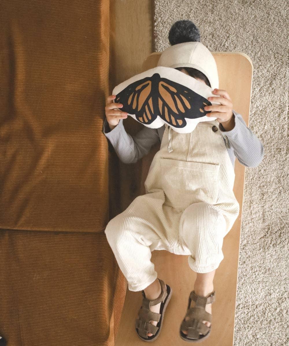 Child in beige overalls and grey shirt lies on wooden bench holding butterfly pillow over face. Come see us at Awaken, Franklin, Tennessee
