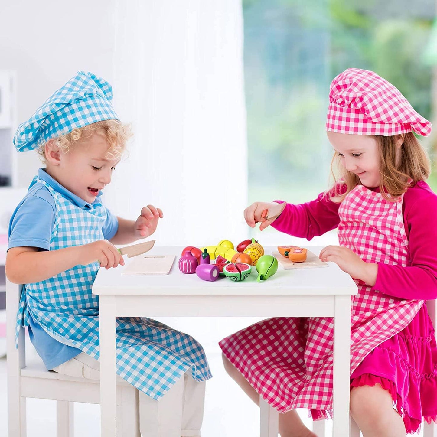 Two kids in chef hats and aprons playing with wooden toy fruits at a white table. Come see us at Awaken, Franklin, Tennessee