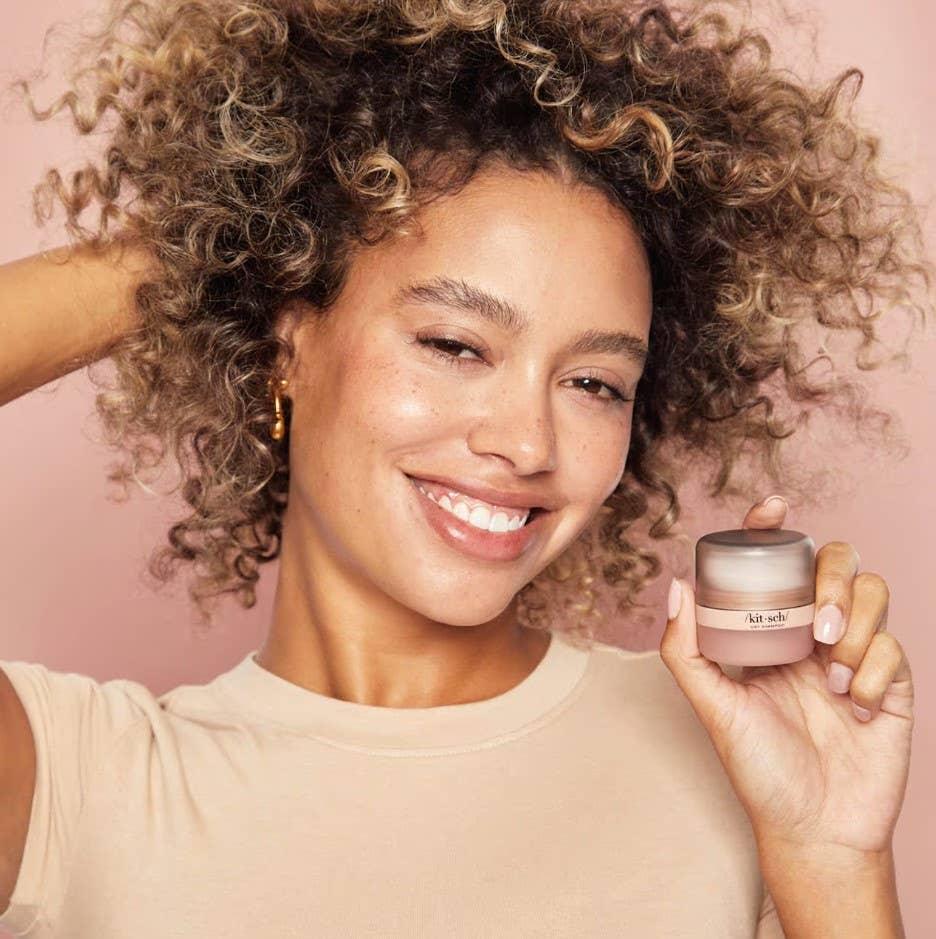 Smiling woman with curly hair holding skincare cream jar, pink background. Come see us at Awaken, Franklin, Tennessee.