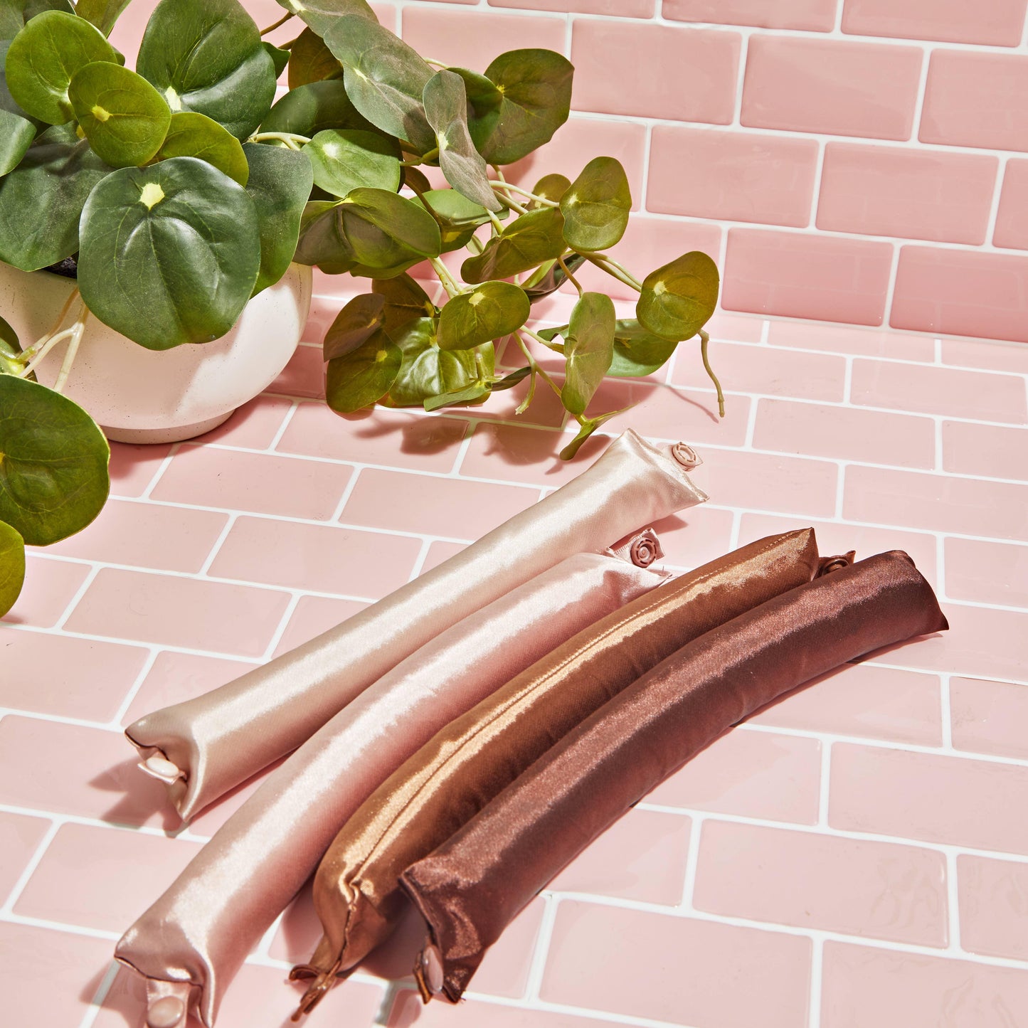 Silky padded hair curlers in nude and brown shades on pink tiles with a green potted plant. Come see us at Awaken, Franklin, Tennessee