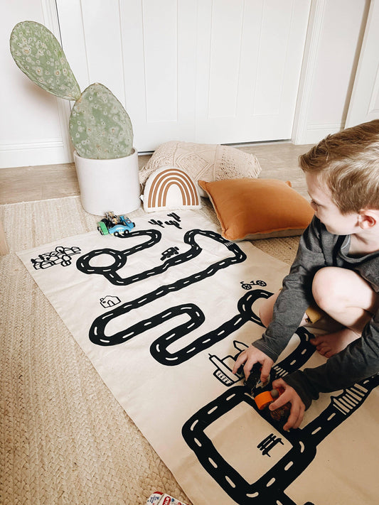 Young boy playing with toy cars on a black and white road play mat near pillows and cactus plant. Come see us at Awaken, Franklin, Tennessee