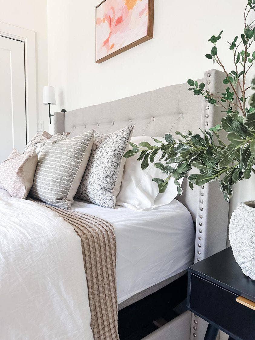 Cozy beige upholstered bed with patterned pillows, textured blanket, and green plant on nightstand. Come see us at Awaken, Franklin, Tennessee