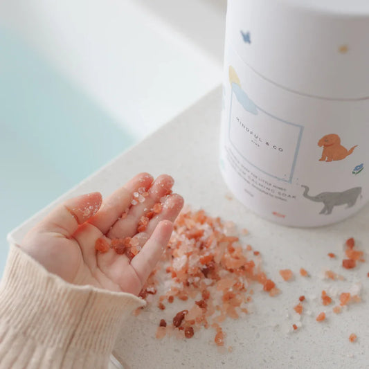 Hand holding pink Himalayan bath salts near Mindful & Co Kids soaking soak container on white surface. Come see us at Awaken, Franklin, Tennessee