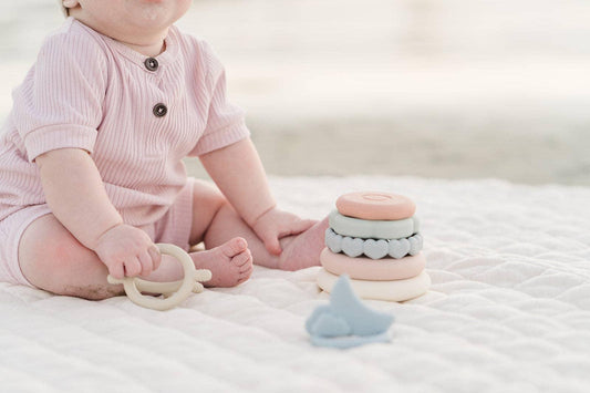 Baby in pink onesie playing with pastel stacking rings and teether on white quilt. Come see us at Awaken, Franklin, Tennessee
