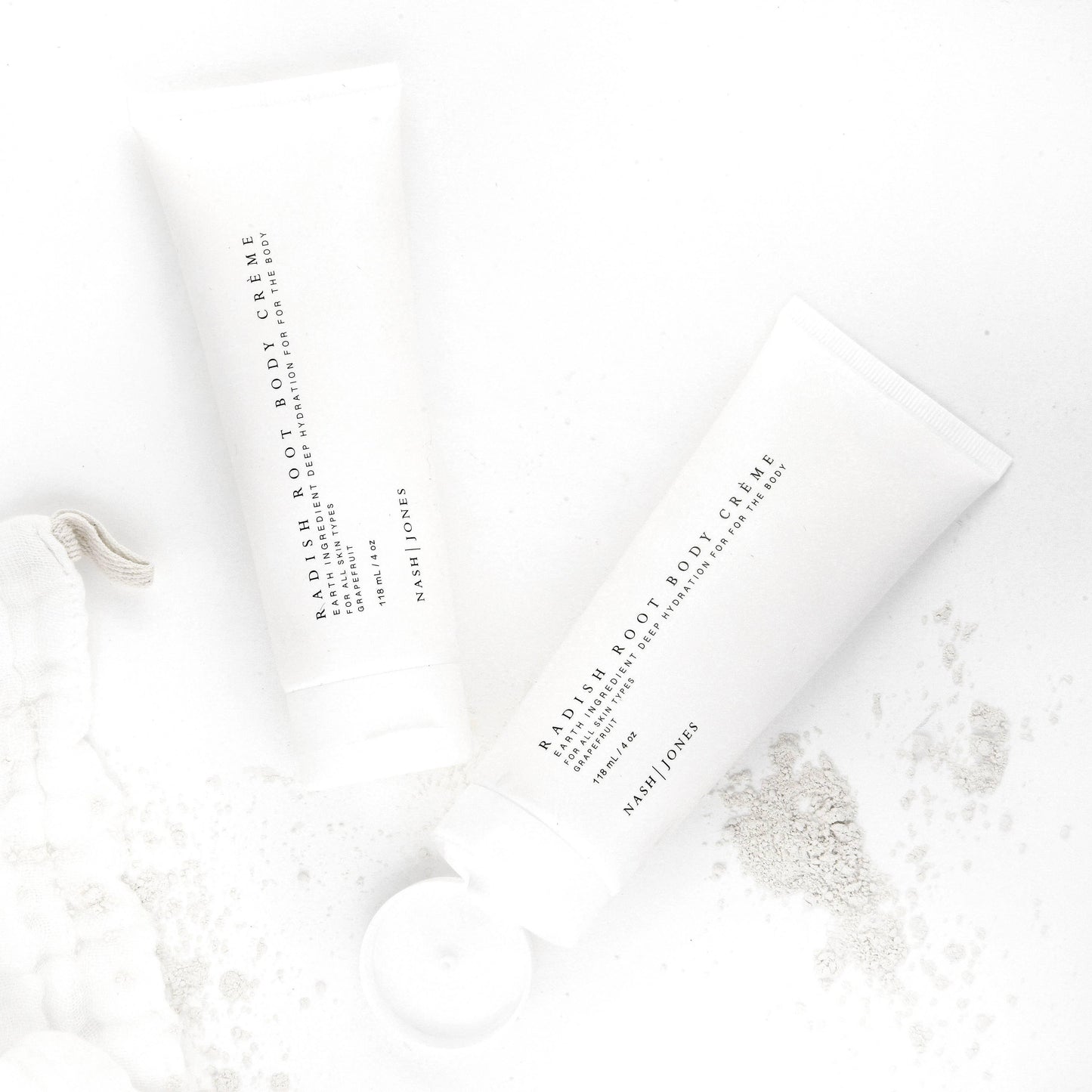 Two tubes of Nash Jones Radish Root Body Crème with grapefruit scent on a white background with powder spills. Come see us at Awaken, Franklin, Tennessee