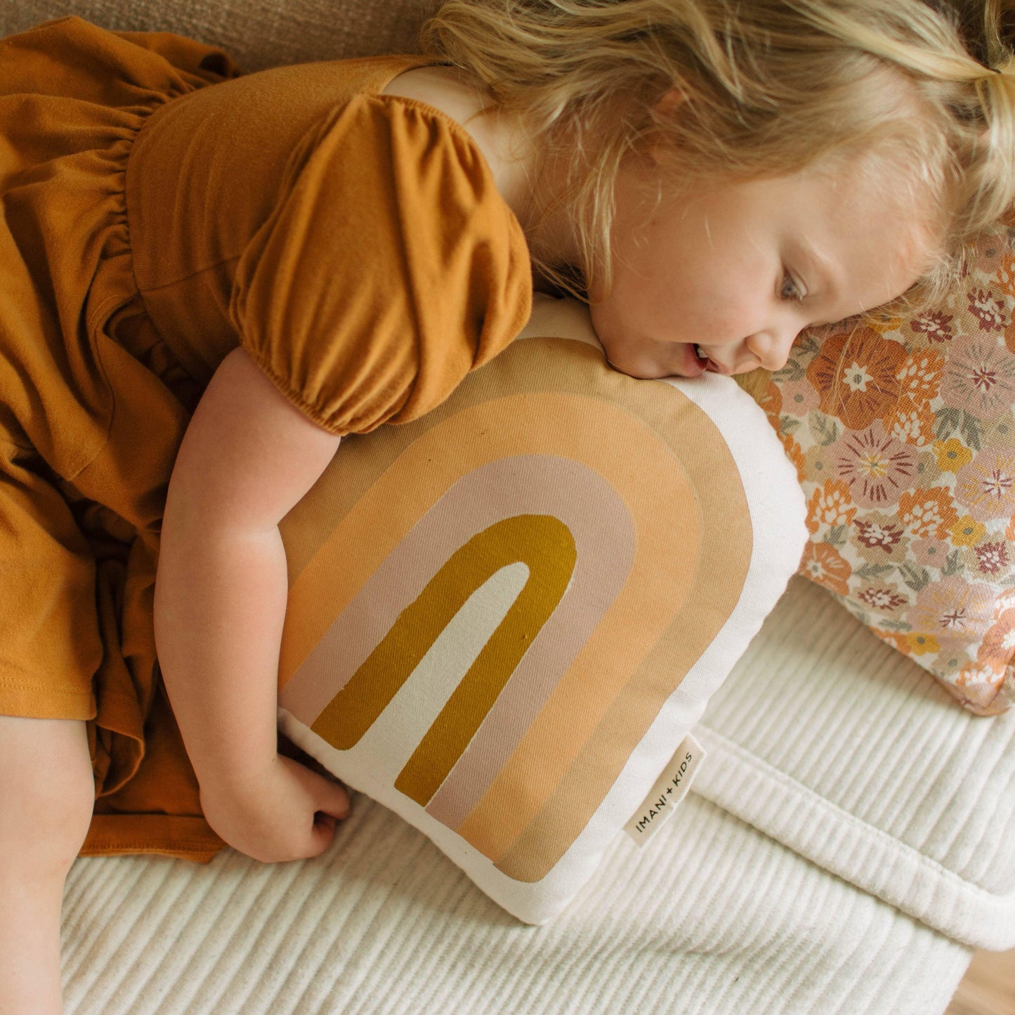 Toddler girl in mustard dress hugging rainbow pillow and floral cushion on white quilted sofa. Come see us at Awaken, Franklin, Tennessee