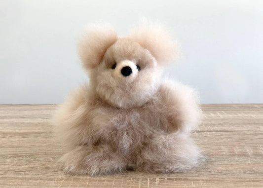 Fluffy beige teddy bear sitting on wooden surface against plain white background. Come see us at Awaken, Franklin, Tennessee