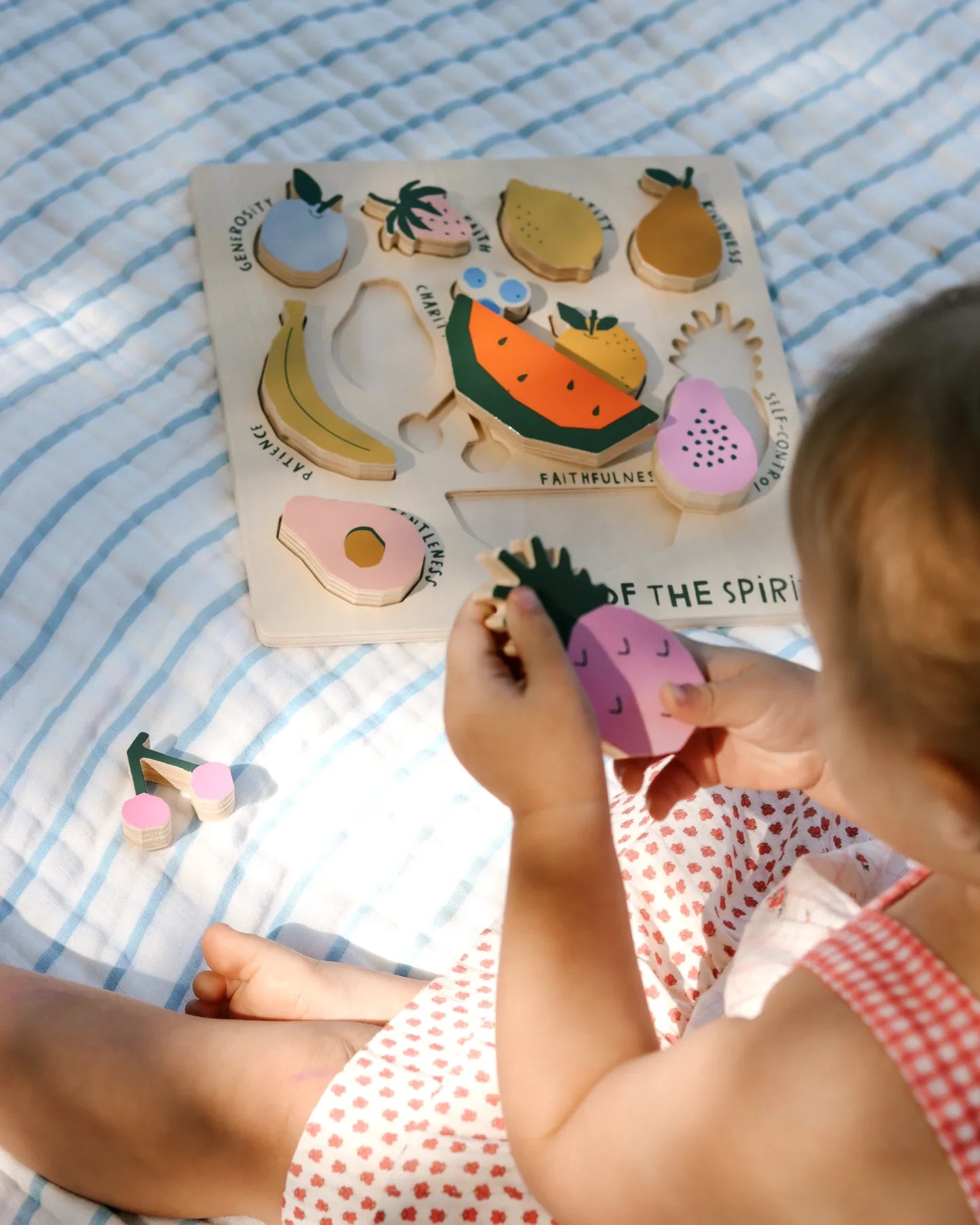 Child playing with wooden fruit puzzle on striped blanket, learning fruits and patience. Come see us at Awaken, Franklin, Tennessee