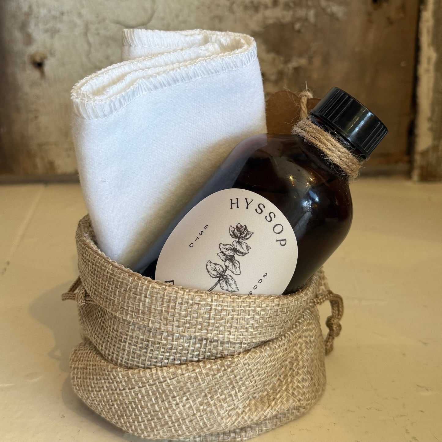 Brown bottle labeled Hyssop inside rustic burlap pouch with white folded cloth, wooden background. Come see us at Awaken, Franklin, Tennessee