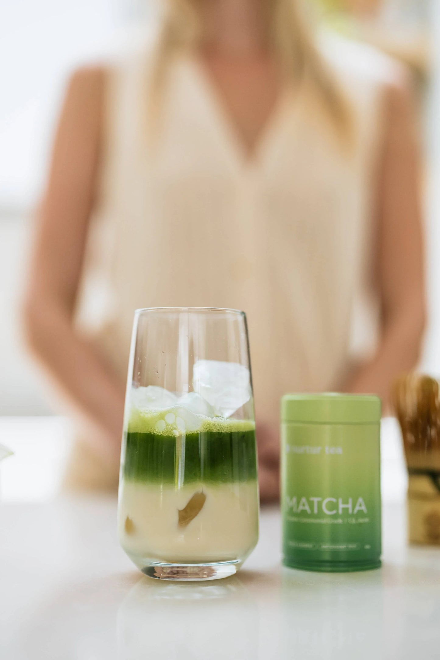 Iced matcha latte in glass with matcha powder container on countertop, blurred person in background. Come see us at Awaken, Franklin, Tennessee