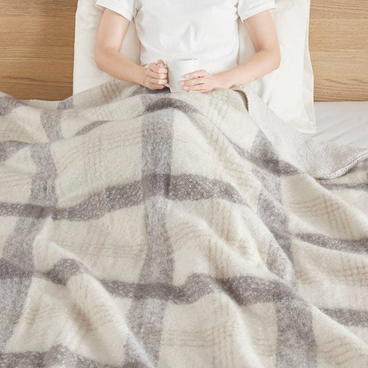 Person in bed holding white mug, covered with soft beige and gray plaid blanket. Come see us at Awaken, Franklin, Tennessee