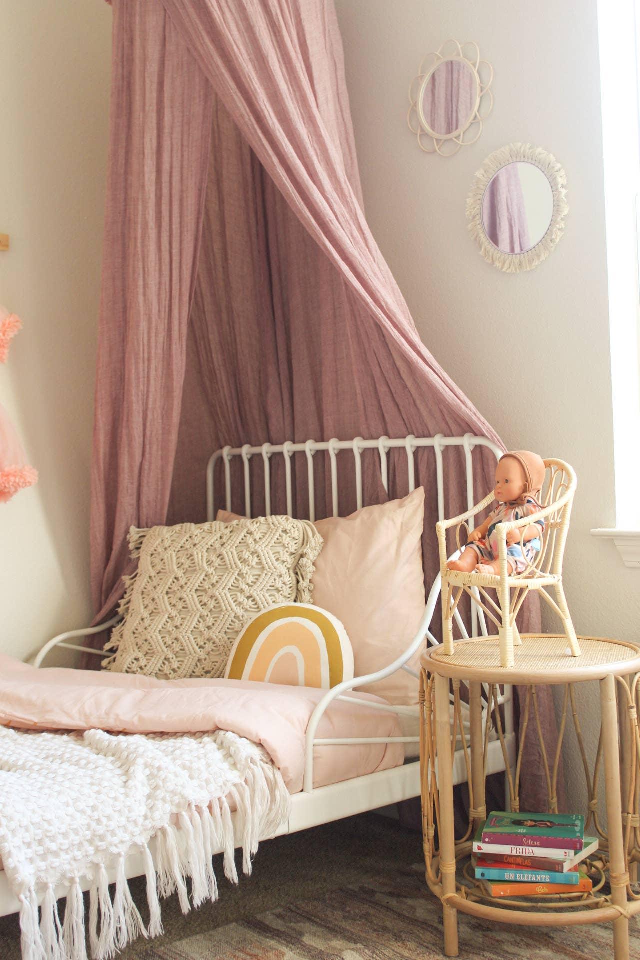 Cozy children's bedroom with white metal bed, pink canopy, patterned pillows, rattan side table and doll. Come see us at Awaken, Franklin, Tennessee