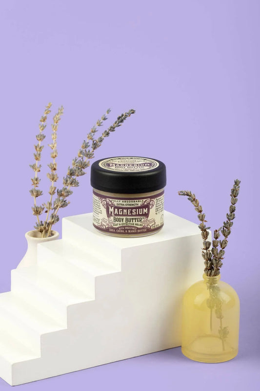 Magnesium body butter jar on white stair-like block with dried lavender in vases, lavender background. Come see us at Awaken, Franklin, Tennessee