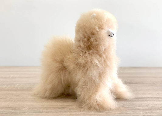 Fluffy beige alpaca figurine on wood surface with plain white background. Come see us at Awaken, Franklin, Tennessee