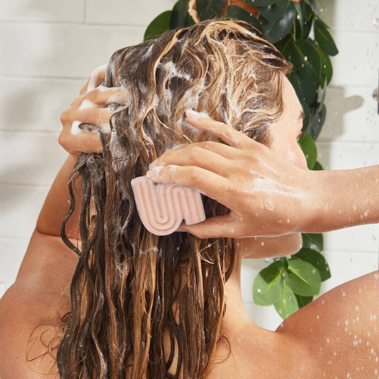 Person washing long wet hair with pink shampoo bar in shower, green plant background. Come see us at Awaken, Franklin, Tennessee