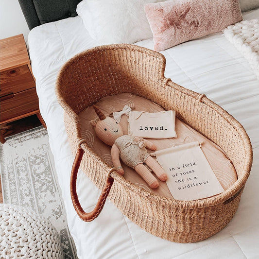 Woven baby bassinet with knitted unicorn toy, baby outfit, and cloth sign on bed - Come see us at Awaken, Franklin, Tennessee