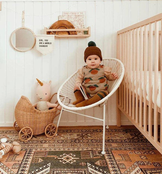 Baby in fall-themed outfit sitting in white chair in cozy nursery with wooden crib and toys. Come see us at Awaken, Franklin, Tennessee