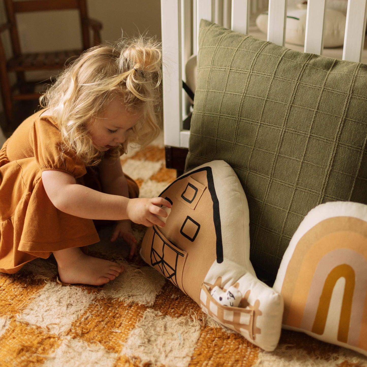 Toddler girl in brown dress playing with plush house and rainbow pillows on rug. Come see us at Awaken, Franklin, Tennessee
