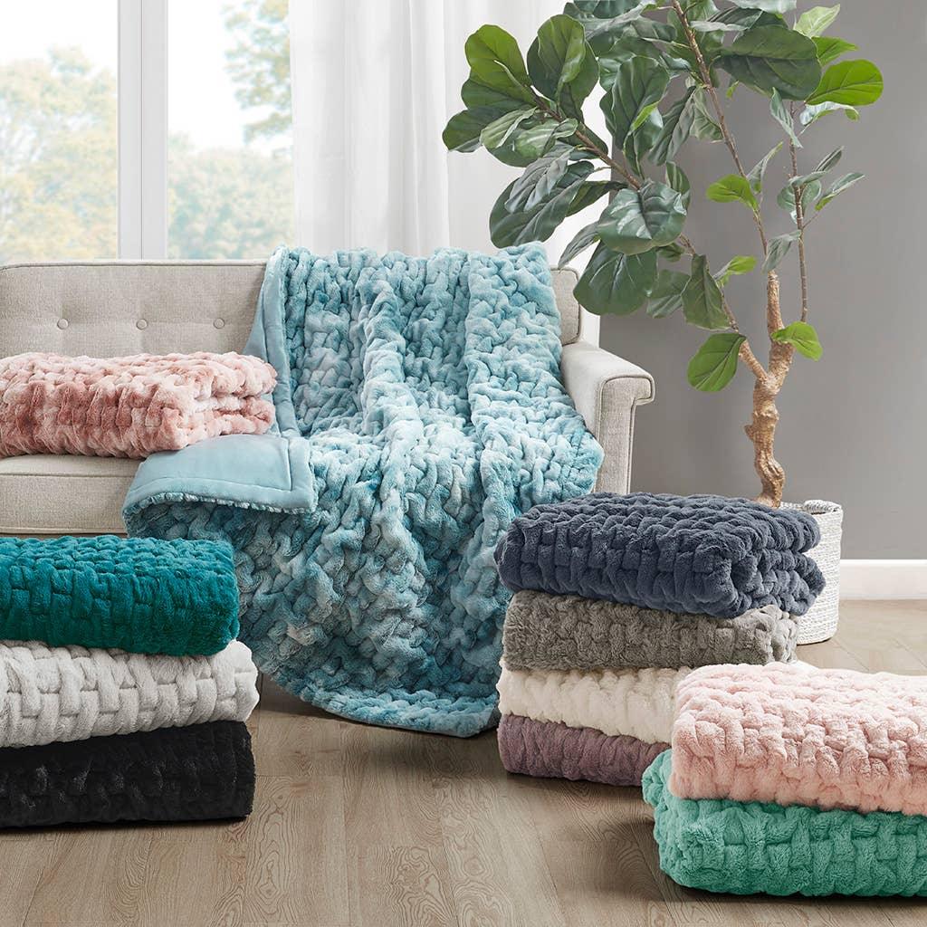 Soft textured blankets in assorted colors displayed on and around a beige sofa with a green indoor plant, cozy living room decor. Come see us at Awaken, Franklin, Tennessee