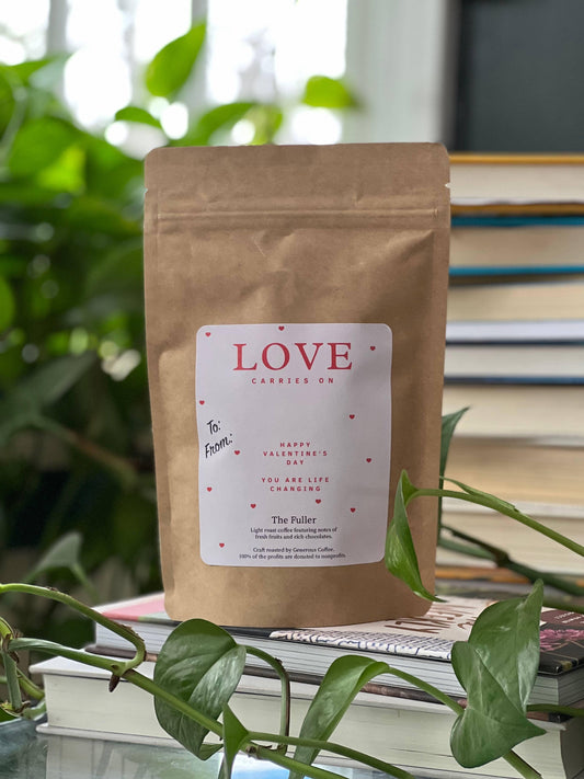 Brown coffee bag labeled Love Carries On on stacked books with green plant in background. Come see us at Awaken, Franklin, Tennessee