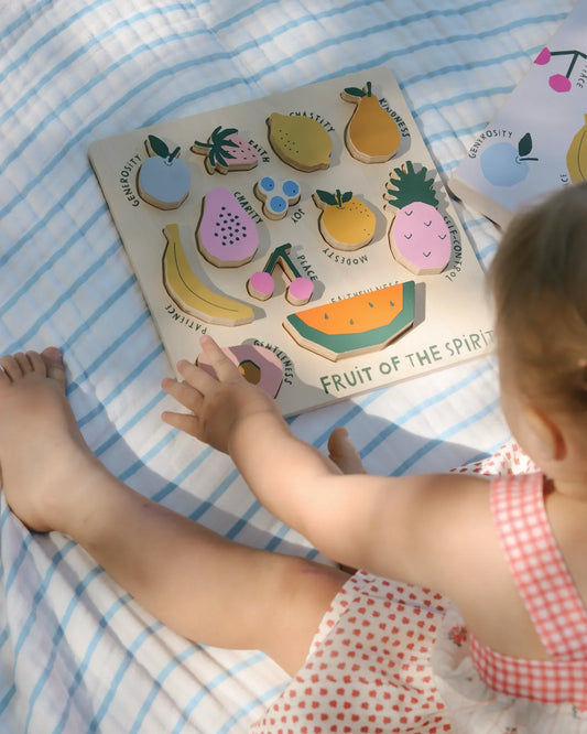 Toddler playing with wooden Fruit of the Spirit puzzle on blue-striped blanket. Come see us at Awaken, Franklin, Tennessee