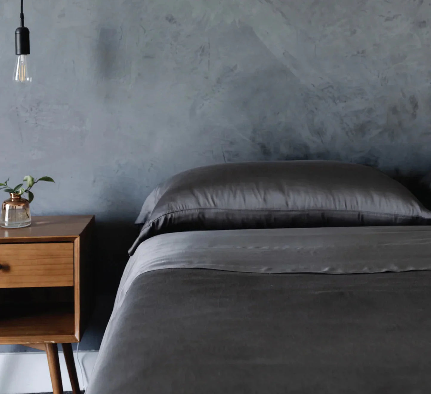 Modern bedroom with dark gray bedding, wooden nightstand with plant, concrete wall, and hanging light bulb. Come see us at Awaken, Franklin, Tennessee