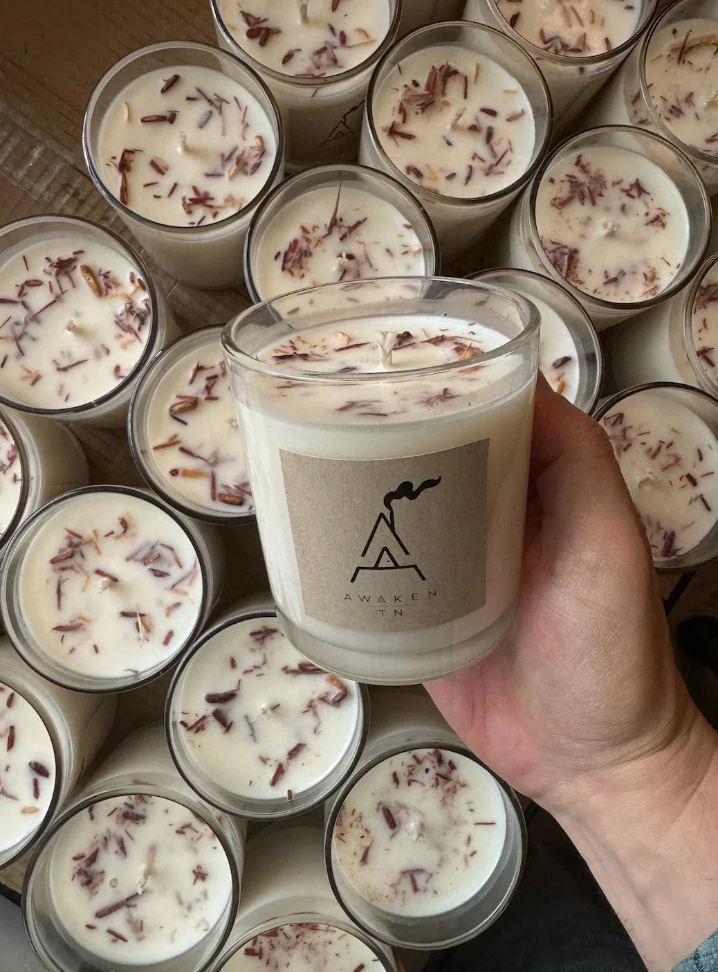 Hand holding Awaken TN scented candle with wood shavings, surrounded by similar candles. Come see us at Awaken, Franklin, Tennessee