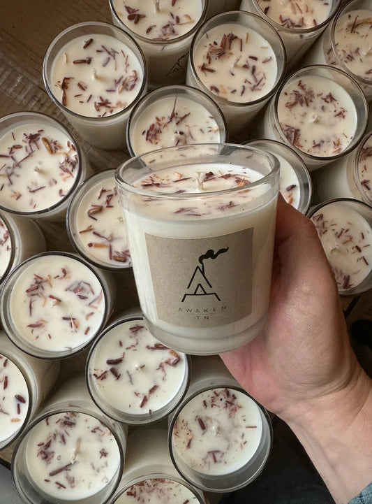 Hand holding Awaken TN scented candle with wood shavings, surrounded by similar candles. Come see us at Awaken, Franklin, Tennessee