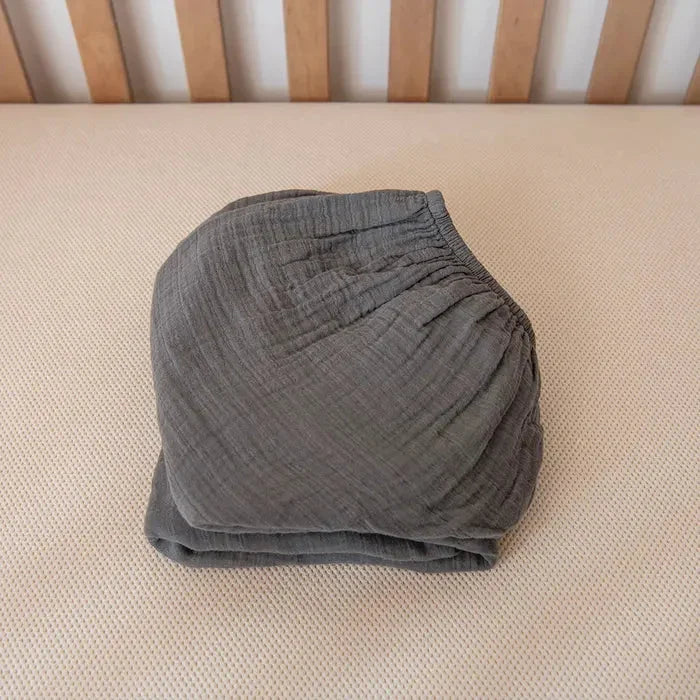 Folded dark gray muslin crib fitted sheet on beige textured crib mattress with wooden slats. Come see us at Awaken, Franklin, Tennessee