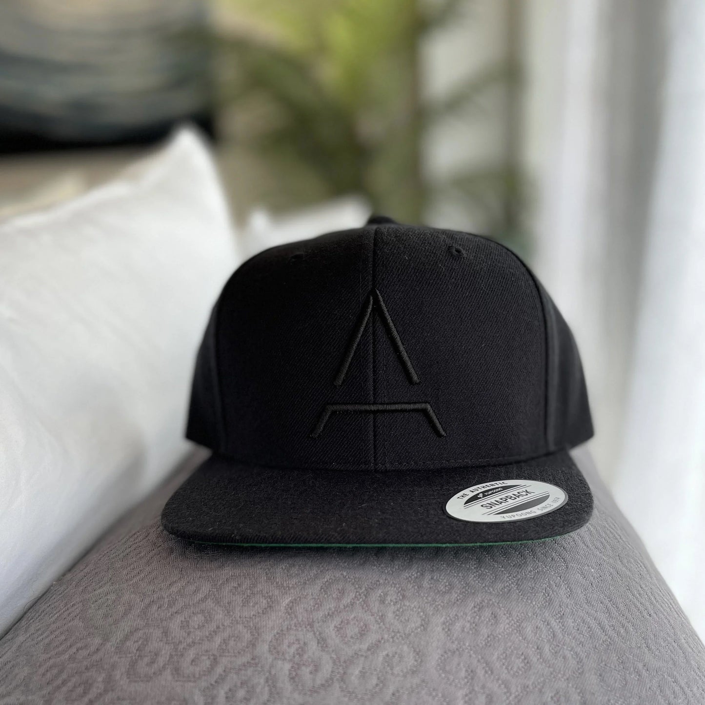 Black snapback hat with embroidered logo on patterned gray surface, blurred cozy background. Come see us at Awaken, Franklin, Tennessee