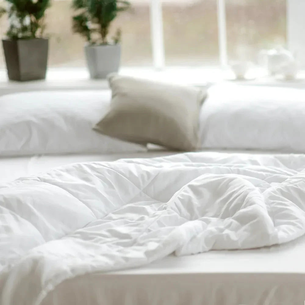 Cozy white bed with comforter and pillow by window with plants Come see us at Awaken, Franklin, Tennessee