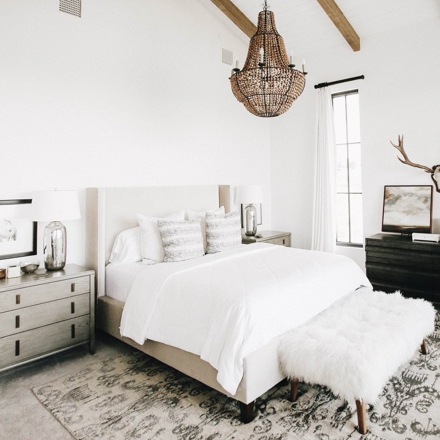 Bright modern bedroom with white bedding, textured pillows, wooden chandelier, fur bench, and rustic decor. Come see us at Awaken, Franklin, Tennessee