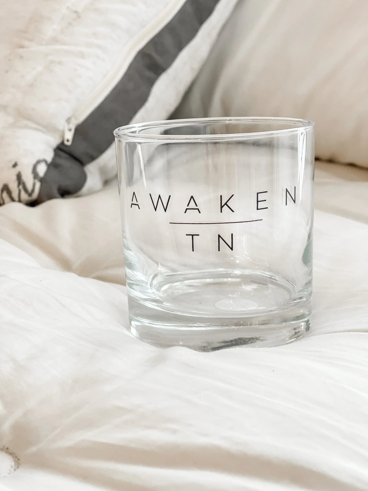 Clear glass with AWAKEN TN logo on white bedding and pillow, cozy home setting. Come see us at Awaken, Franklin, Tennessee