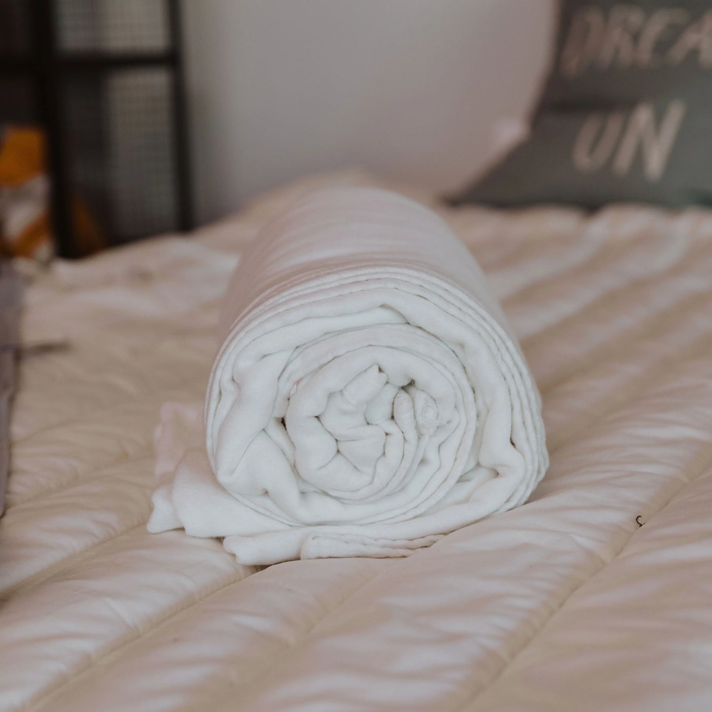 Rolled white blanket on a bed with quilted bedding and decorative pillows, cozy bedroom scene. Come see us at Awaken, Franklin, Tennessee