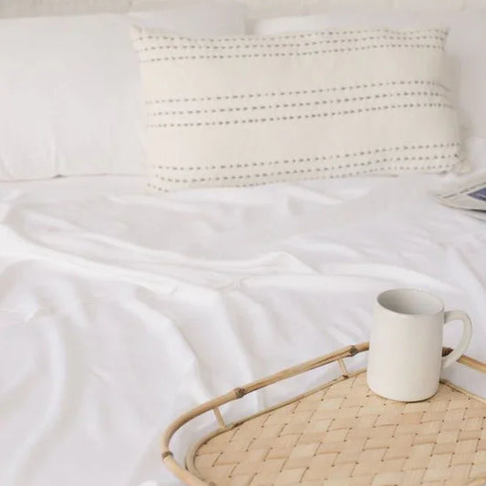 White ceramic mug on woven tray placed on white bed linen with stitched pillow in background. Come see us at Awaken, Franklin, Tennessee